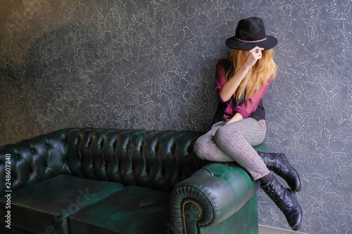 Canvas Print Girl hipster in a black bowler hat sitting on a leather green sofa, covering her face with a hat