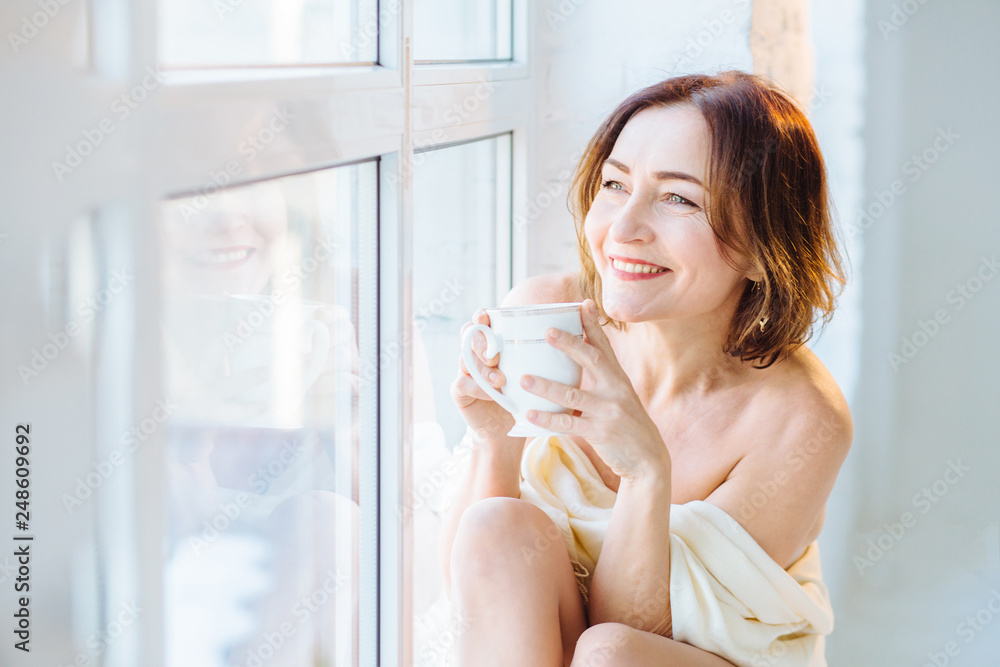 Beautiful happy miling mature woman with clear skin and cute smile. Female sitting on windowsill drinking tea. Elegant, vulnerable, fond, gentle concept. Spa and skincare. Beautiful happy miling mature woman with clear skin and cute smile. Female sitting on windowsill drinking tea. Elegant, vulnerable, fond, gentle concept. Spa and skincare.