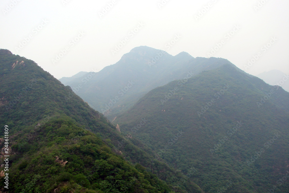 Panorama of mountain ridges covered with dense forest through the prism of a foggy day from the height of the stairs of the Great Chinese wall against the background of the cloudy sky.