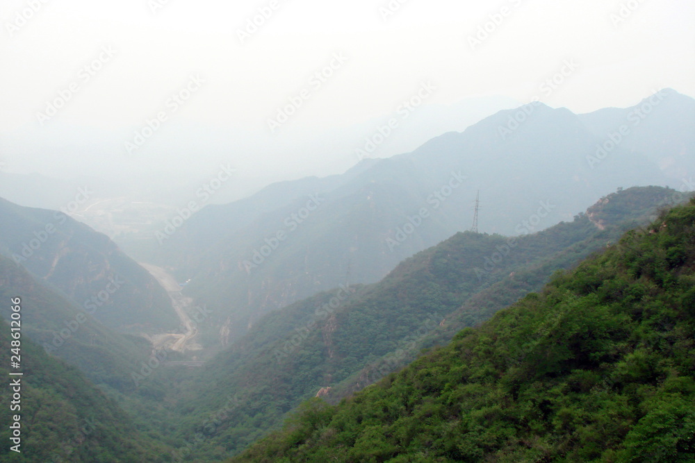 Panorama of mountain ridges covered with dense forest through the prism of a foggy day from the height of the stairs of the Great Chinese wall against the background of the cloudy sky.