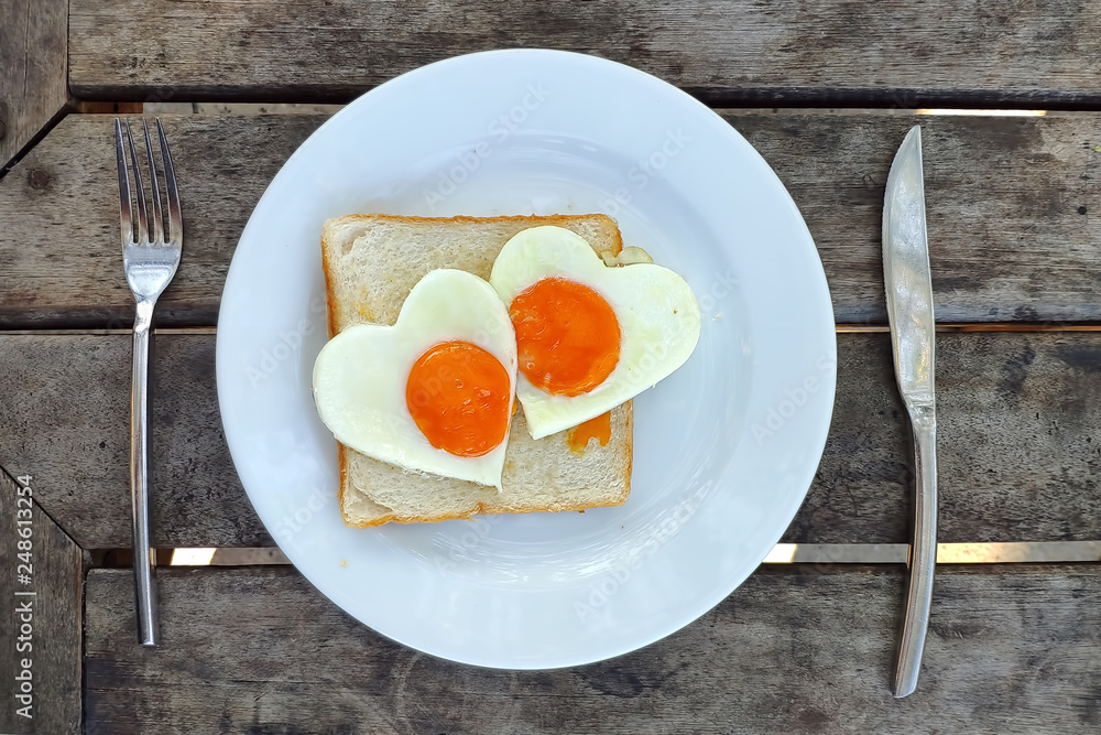 Breakfast for Valentines day. Fried eggs in the shape of a heart at toast on white plate and wooden table. Knife and fork lie nearby Breakfast for Valentines day. Fried eggs in the shape of a heart at toast on white plate and wooden table. Knife and fork lie nearby