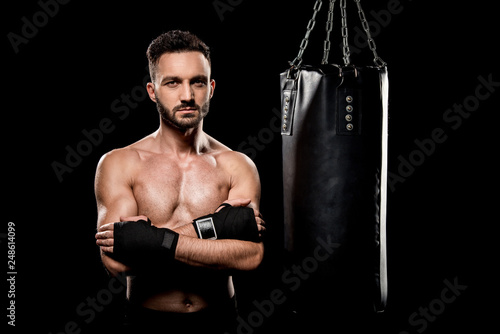 muscular boxer standing with crossed arms near punching bag isolated on black
