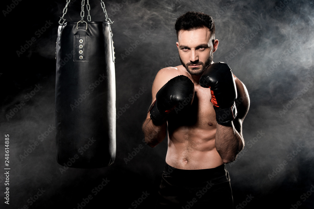 muscular athlete standing in boxing pose near punching bag on black with smoke