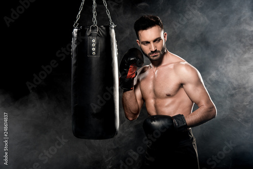 muscular sportsman in boxing gloves standing near punching bag on black with smoke