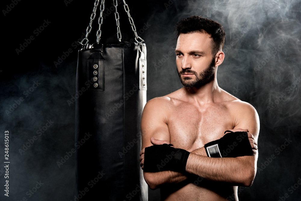 thoughtful man standing with crossed arms near punching bag on black with smoke thoughtful man standing with crossed arms near punching bag on black with smoke