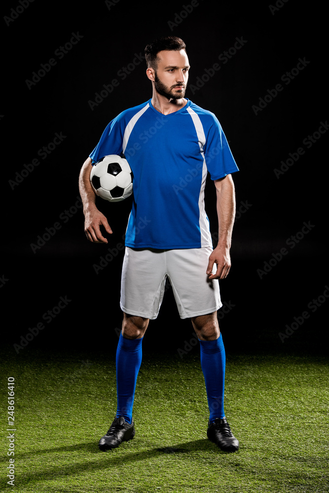 handsome football player standing with ball on green grass isolated on black