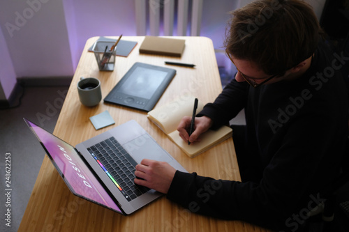 Young Handsome Graphic Designer Illustrator Man at Work. Black Sweatshirt. Cozy Workplace Wooden Table, Laptop, Tablet Pen, Coffee Mug, Sketchbook. Creative Design Working Process. Dark Atmosphere.