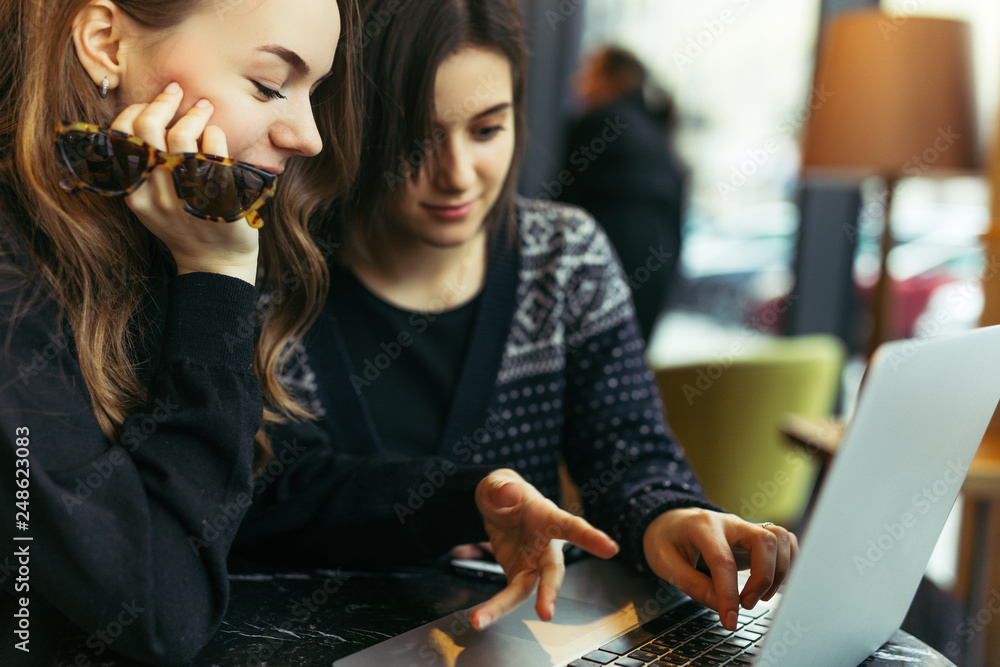 Fototapeta premium Two girls, friends Drink Hot Coffee While Work In Cafe On Laptop. Portrait Of Stylish Smiling Girls In Winter Clothes Drinking Hot Coffee And Work At Laptop. Female Winter Style. - Image