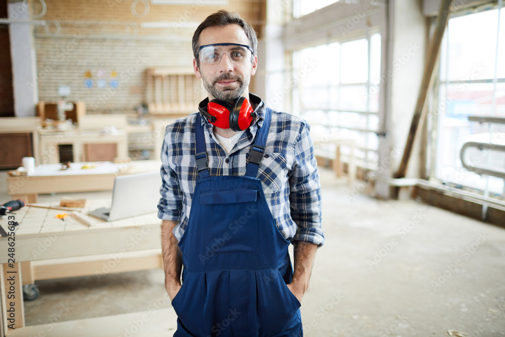 Content satisfied handsome middle-aged carpenter in safety goggles wearing overall and ear protectors on neck holding hands in pockets and looking at camera in workshop. Content satisfied handsome middle-aged carpenter in safety goggles wearing overall and ear protectors on neck holding hands in pockets and looking at camera in workshop.