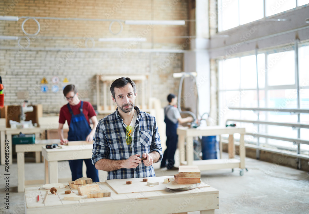 Content handsome middle-aged carpenter in checkered shirt standing at table with wooden details and looking at camera while assembling kids toys Content handsome middle-aged carpenter in checkered shirt standing at table with wooden details and looking at camera while assembling kids toys