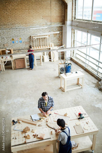 Top view of busy carpenters standing at table and making wooden details for toy assembling, they working with small wooden pieces and chatting in workshop