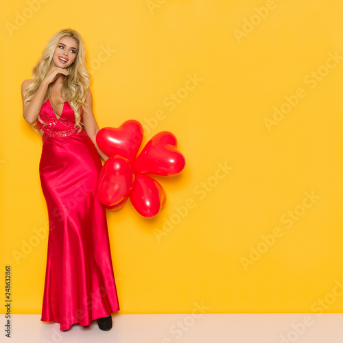 Smiling Beautiful Woman In Elegant Red Dress Is Holding Heart Shaped Balloons And Looking Away