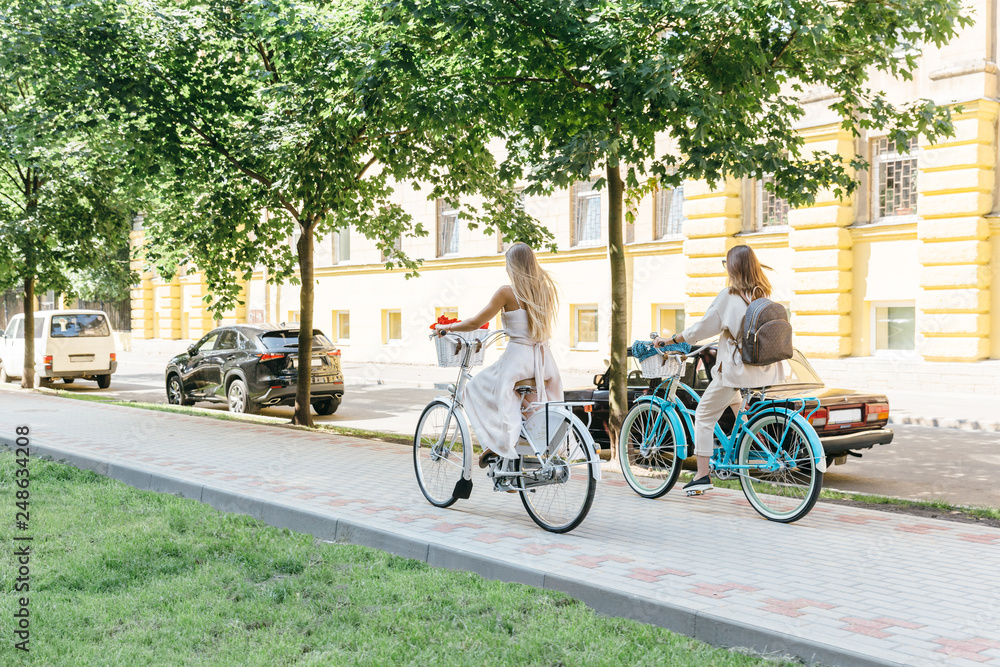 Fototapeta premium Two blonde friends ride on bicycles along the boulevard/ white and blue retro bikes with baskets, sensuality and elegance, healthy and happy cyclist women/ summertime, leisure and lifestyle concept.