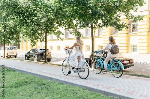 Two blonde friends ride on bicycles along the boulevard/ white and blue retro bikes with baskets, sensuality and elegance, healthy and happy cyclist women/ summertime, leisure and lifestyle concept.