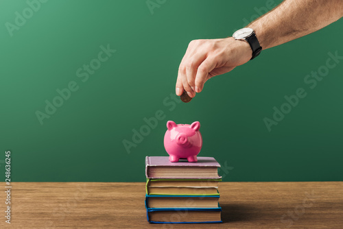 man puting in pink piggy bank coin on books, wooden table and green background photo