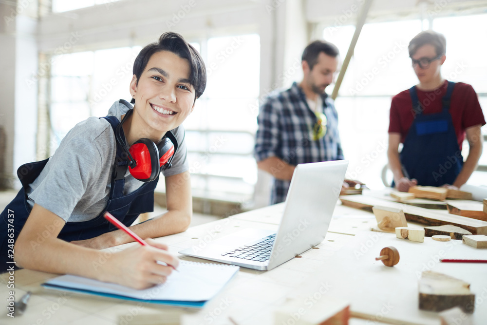 Fototapeta premium Cheerful excited young lady with ear protectors on neck leaning on table and smiling at camera while enjoying work in carpentry