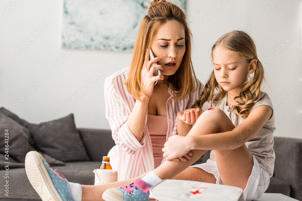 Upset mother talking on smartphone and kid with wound sitting on table at home