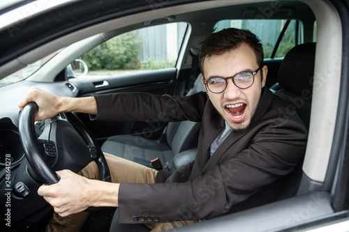 Furious young man is looking at the camera while sitting at his car. He is screaming at someone. His hands are on the steering wheel. Angry driver concept. © Maksym