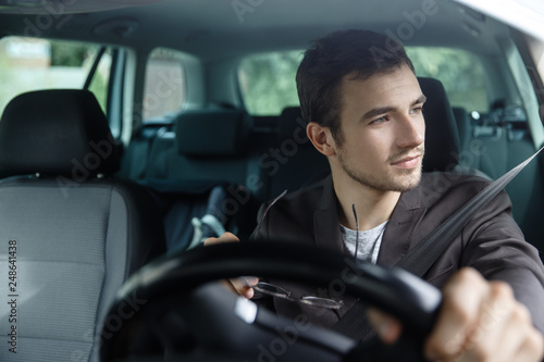 Delighted young man is looking trough the window while driving his car. He is holding his glasses at his right hand. His left hand is on the steering wheel.