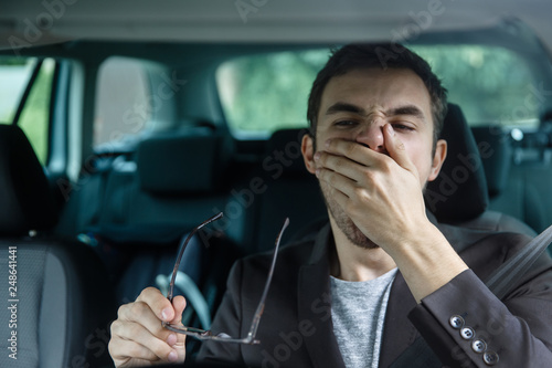 Sleepy young guy yawns while covering his mouth with his left hand. He is holding glasses at his right hand. Road safety concept.