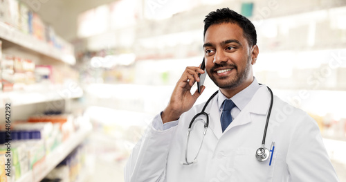 medicine, technology and healthcare concept - smiling indian male doctor or pharmacist in white coat with stethoscope calling on smartphone over drugstore background photo