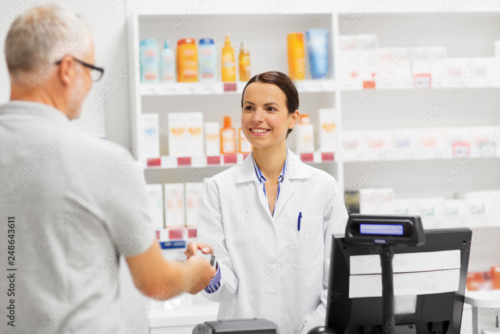 medicine, pharmaceutics, healthcare and people concept - happy smiling female apothecary taking senior customer credit card at pharmacy cash register