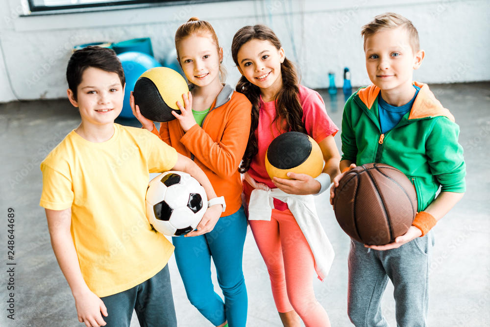 Excited children in sportswear posing with balls - obrazy, fototapety, plakaty 