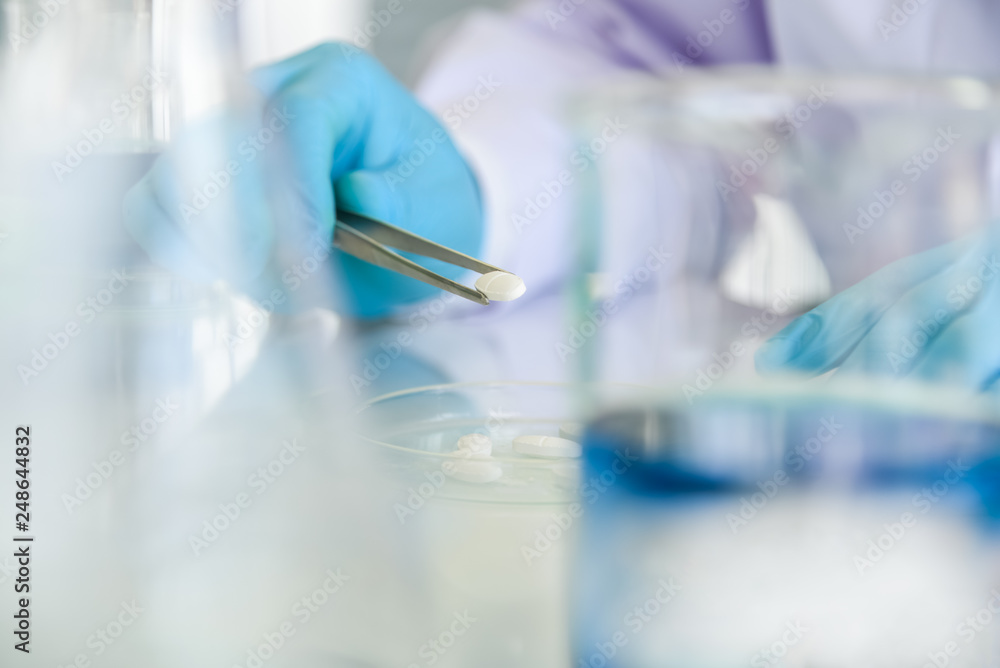 Blurred soft images of Scientists use clamps to clamp the white pill tablests, from glass cups for inspection and research, to science concept. Blurred soft images of Scientists use clamps to clamp the white pill tablests, from glass cups for inspection and research, to science concept.