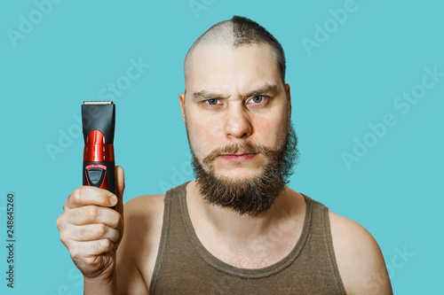 Portrait of a bearded uncut man, who himself cuts his beard, hair on his head and mustache with a clipper at home on an isolated colored background. Concept before after photo