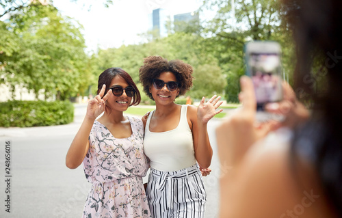 female friendship, technology and people - woman with smartphone photographing her friends showing peace hand sign in summer park