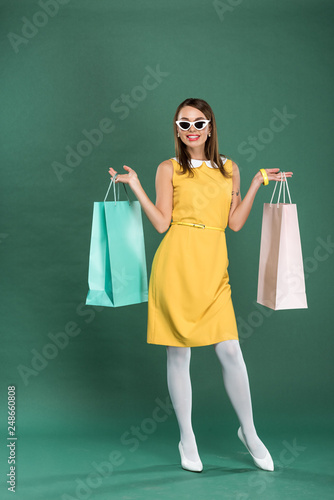 smiling stylish woman in yellow dress and sunglasses with shopping bags posing on green background © LIGHTFIELD STUDIOS