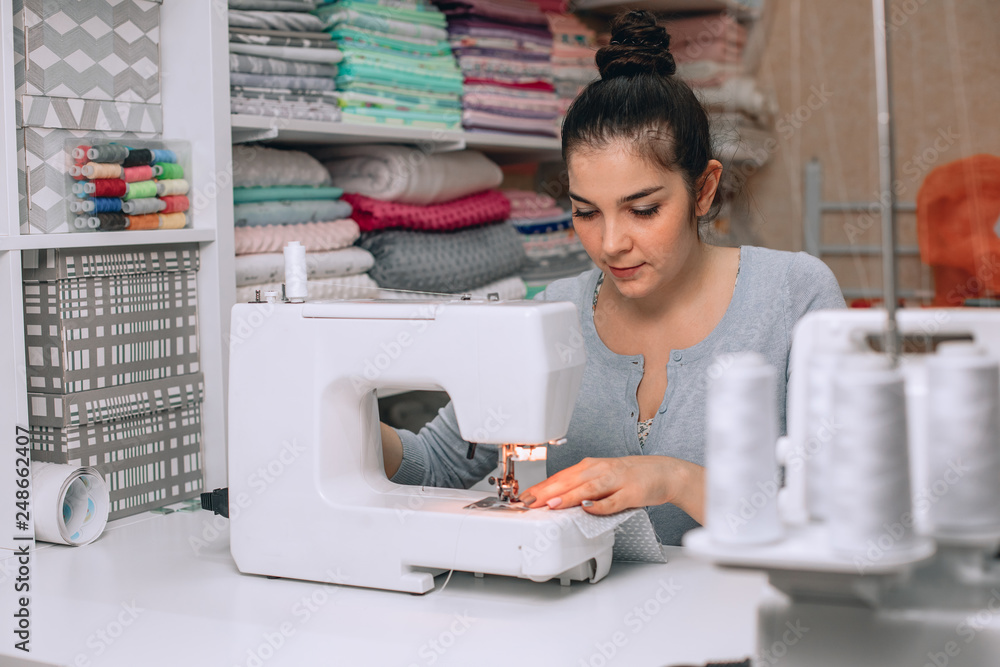 portrait of young woman seamstress sitting and sews on sewing machine. Tailor making a garment in her workplace. Hobby as a small business concept. portrait of young woman seamstress sitting and sews on sewing machine. Tailor making a garment in her workplace. Hobby as a small business concept.