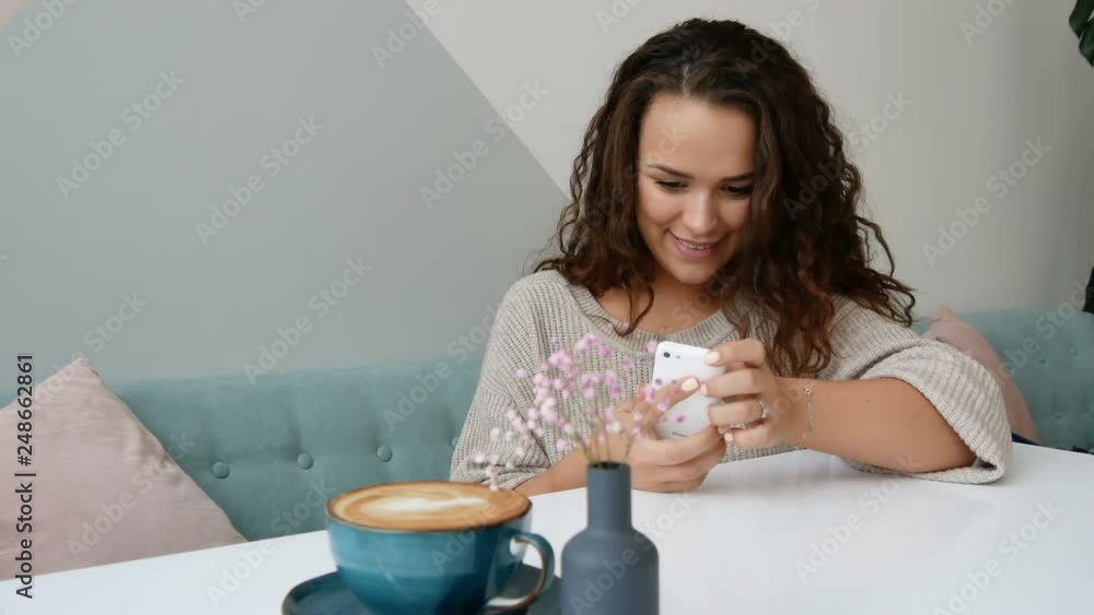 Attractive woman take mobile photo of cappuccino at cafe. Curly brunette use smartphone at modern cafe. Attractive woman take mobile photo of cappuccino at cafe. Curly brunette use smartphone at modern cafe.