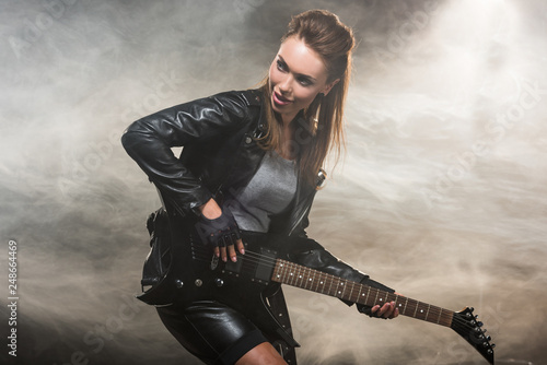 beautiful woman in leather jacket playing electric guitar on smoky background