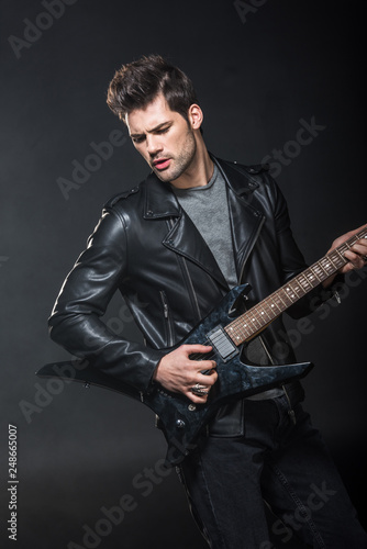 handsome rocker in leather jacket playing electric guitar isolated on black