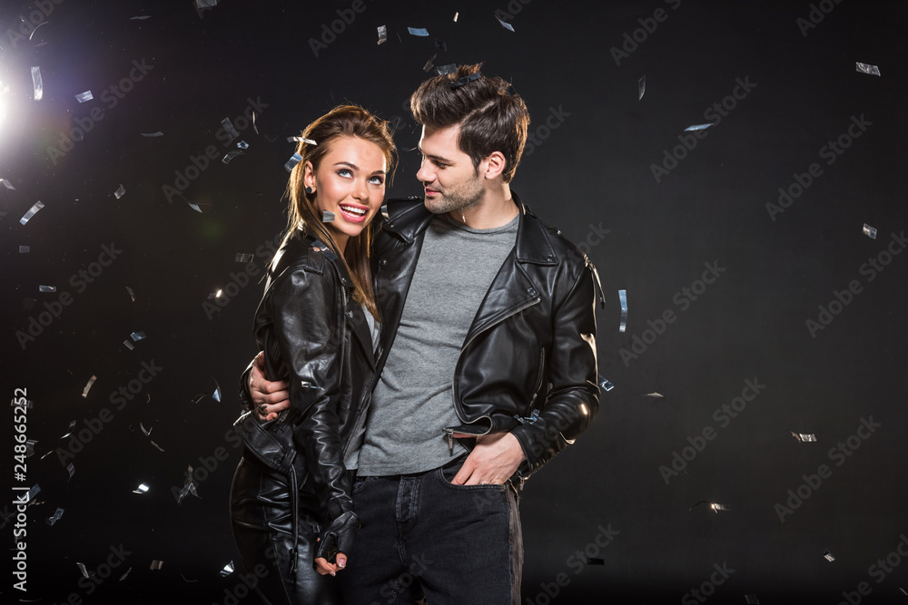 beautiful couple in leather jackets hugging with falling confetti on black background