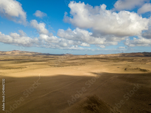 Fototapeta Naklejka Na Ścianę i Meble -  Aerial view of a desert landscape on the island of Lanzarote, Canary Islands, Spain. Road that crosses a desert. Tongue of black asphalt cutting a desert land. Reliefs on the horizon. Volcanoes