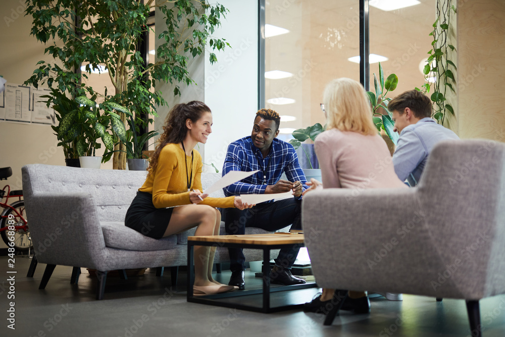 Positive creative business team in casual clothing sitting on sofas and discussing sales statistics while analyzing papers in office Positive creative business team in casual clothing sitting on sofas and discussing sales statistics while analyzing papers in office
