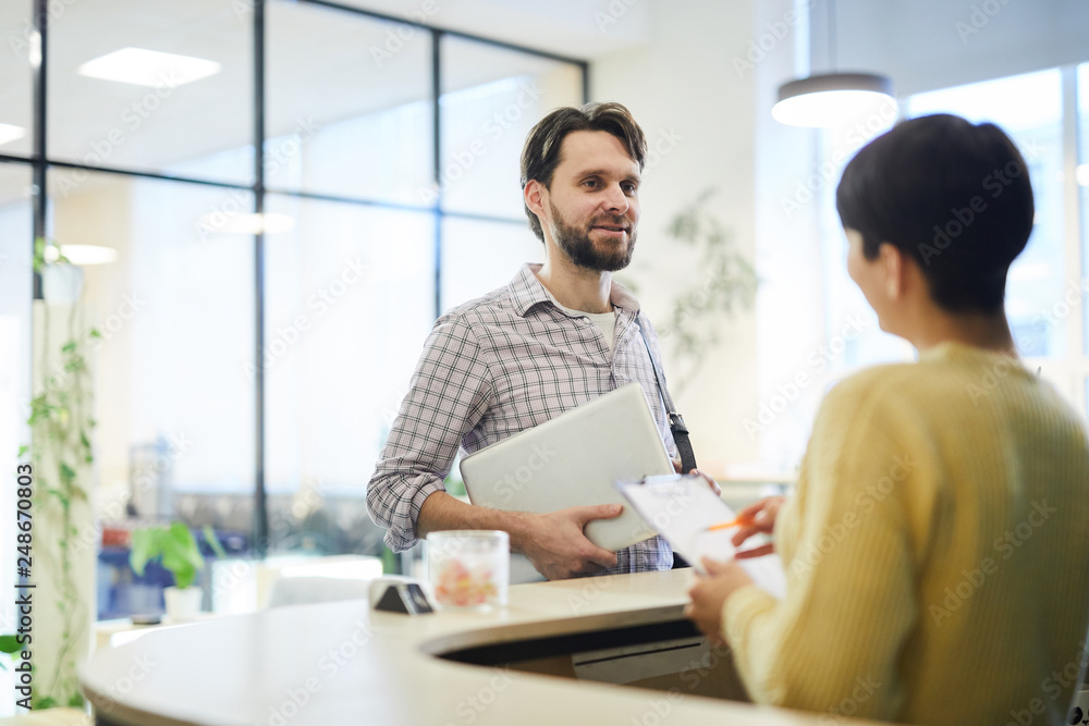 Content handsome young programmer with beard standing at reception and holding laptop while talking about accommodation to hotel receptionist Content handsome young programmer with beard standing at reception and holding laptop while talking about accommodation to hotel receptionist