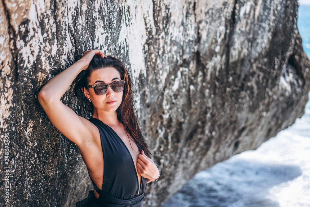 Pretty brunette pan asian girl in black swimsuit relaxing under the rock on the beach.