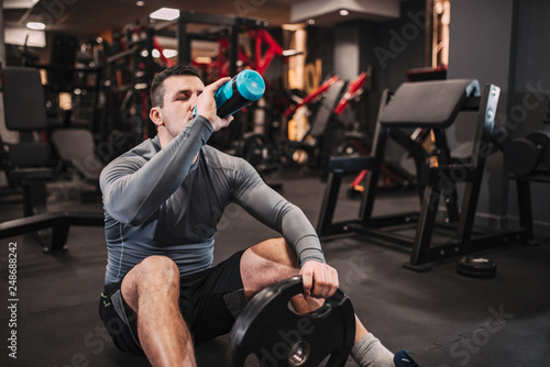 Muscular man drinking water while sitting on the floor in the gym.