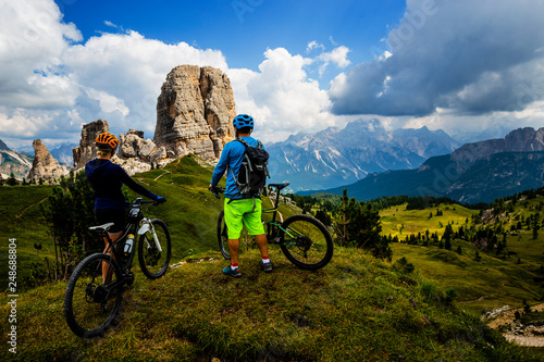 Couple cycling in Cortina d'Ampezzo, stunning Cinque Torri and Tofana in background. Woman and man riding MTB trail. South Tyrol province of Italy, Dolomites.