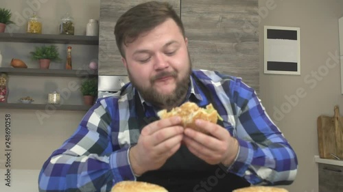 Fat man with a beard gorges on the kitchen fast food hamburgers, extra calories, slow motion, gluttony photo