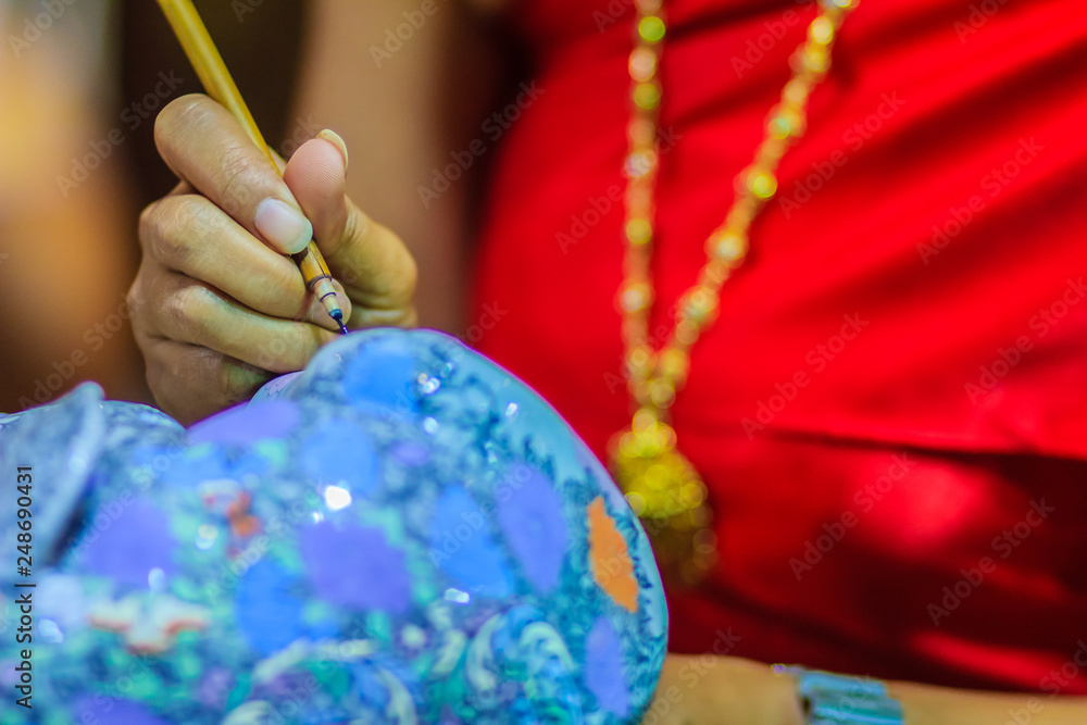 Fototapeta premium Close up Thai woman artist in red Thai style dressed is painting the masterpiece of Benjarong pattern, the famous 5- colored porcelain ceramic wares.