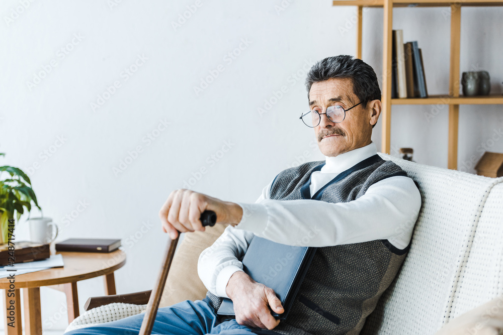 retired man in glasses sitting on sofa with walking stick