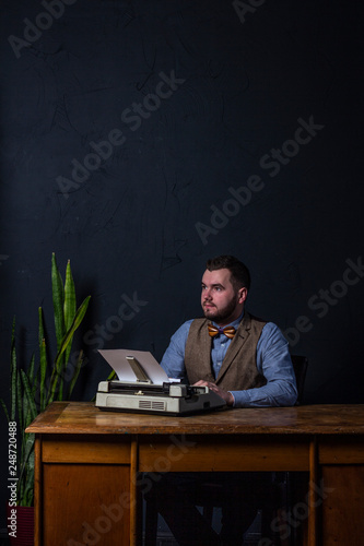 Bearded stylish writer typing on typewriter. Modern writer working on new book in office. Dark background. Space for text