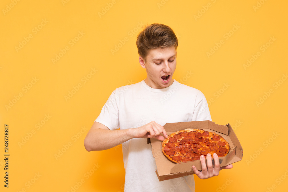 Positive young man in a white T-shirt stands on a yellow background and looks surprised at the box of pizza in his hands. Emotional guy with a box of pizza is isolated on a yellow background. Positive young man in a white T-shirt stands on a yellow background and looks surprised at the box of pizza in his hands. Emotional guy with a box of pizza is isolated on a yellow background.