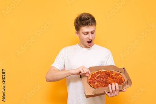 Positive young man in a white T-shirt stands on a yellow background and looks surprised at the box of pizza in his hands. Emotional guy with a box of pizza is isolated on a yellow background.