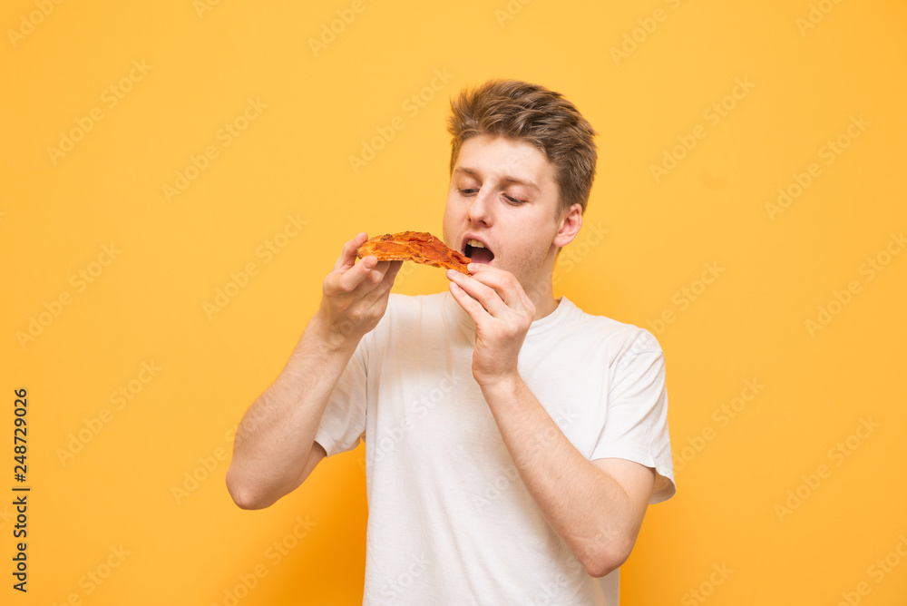 Portrait of a guy in a white T-shirt holds a piece of pizza in his hands, looking and eating. Hungry guy eats fast food, pizza on a yellow background. Copyspace Portrait of a guy in a white T-shirt holds a piece of pizza in his hands, looking and eating. Hungry guy eats fast food, pizza on a yellow background. Copyspace