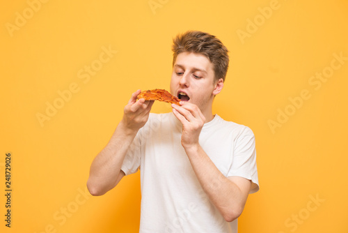 Portrait of a guy in a white T-shirt holds a piece of pizza in his hands  looking and eating. Hungry guy eats fast food  pizza on a yellow background. Copyspace